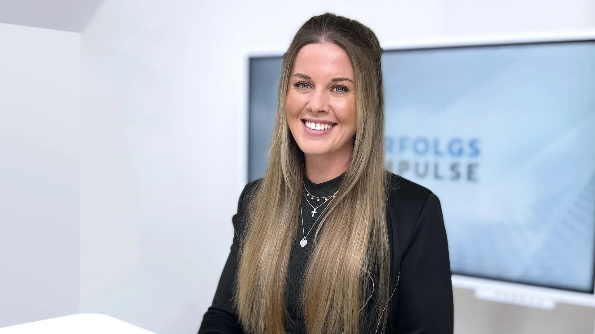 Porträt von Alina Klejbor in schwarzem Blazer im Büro von Erfolgsimpulse, freundlich lächelnd vor einem Bildschirm mit Erfolgsimpulse Schriftzug, als Business Motiv für Coaching, Führung, Persönlichkeitsentwicklung und professionelle Unternehmenskommunikation.