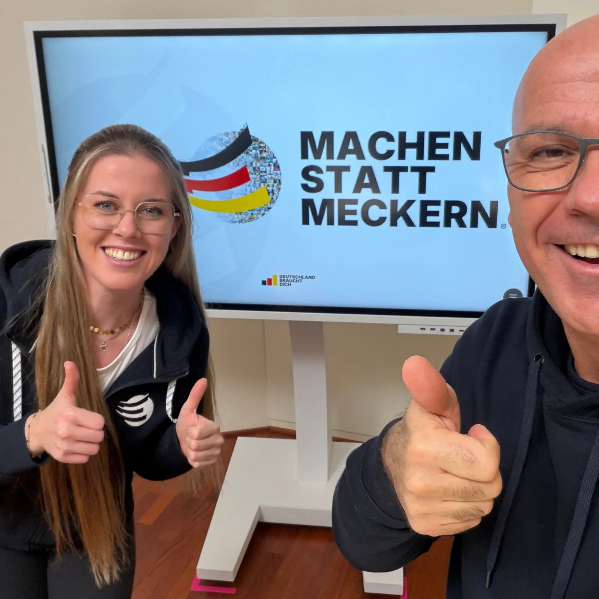 Alina Klejbor links und Marc Klejbor rechts, lächelnd mit Daumen nach oben vor einem Bildschirm mit der Initiative Machen statt Meckern® im Erfolgsimpulse Studio.