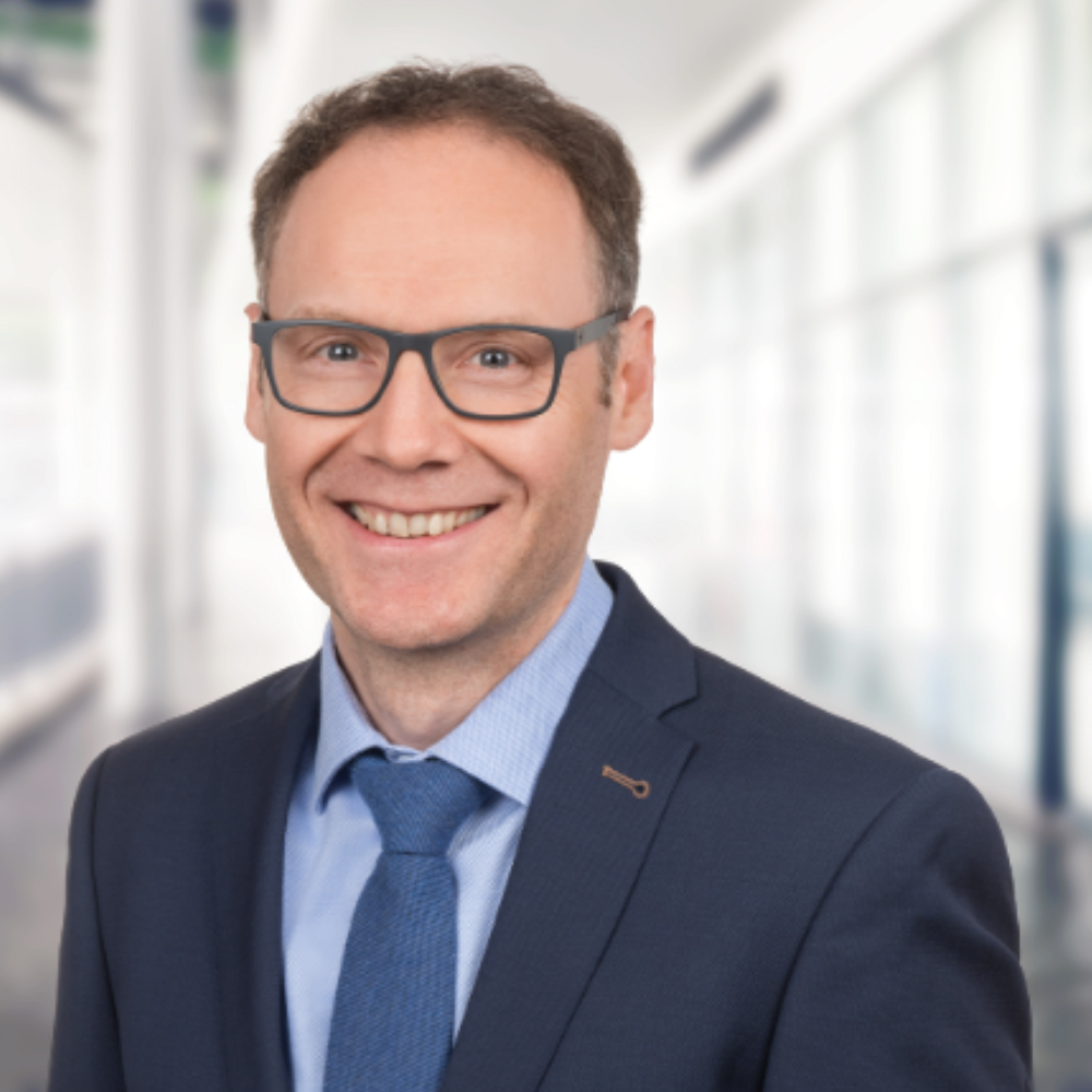 Porträt von Frank Körfer, Steuerberater, im Anzug mit Krawatte vor modernem Hintergrund.