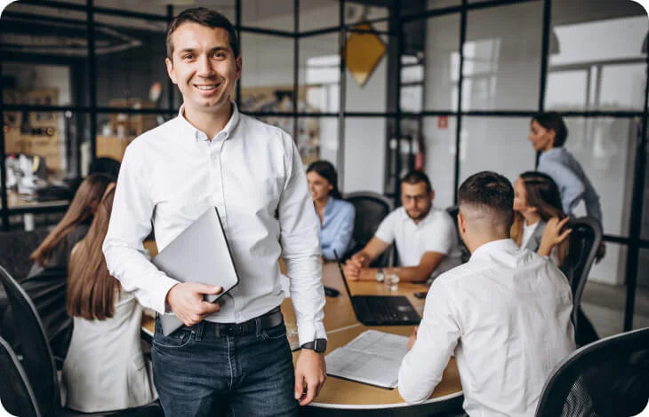 Mitarbeiter mit Laptop steht selbstbewusst vor einem Team Meeting im Büro, Symbolbild für Führung, Zusammenarbeit und professionelle Teamkoordination im Unternehmen.